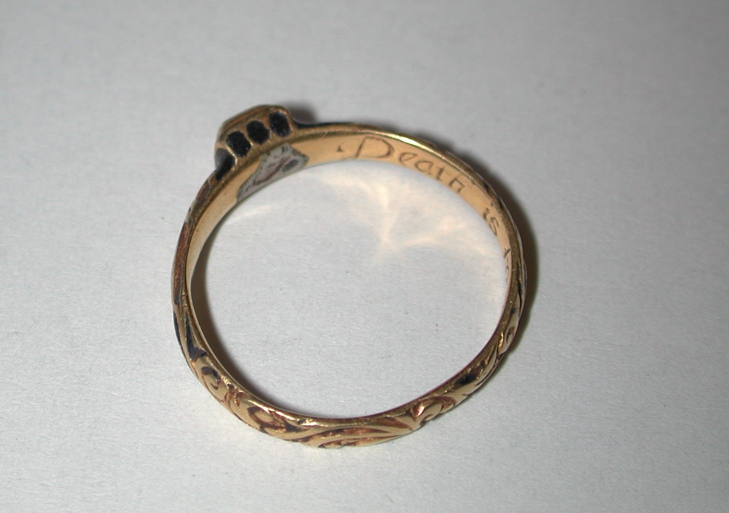 A gold ring seen from the inside, with a script inscription engraved on the inner band and black enamel and engraved leaf patterns on the outside of the hoop