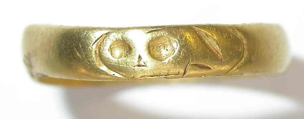 Close view of the bezel of a gold finger ring with a stamped design of brackets and dots that reads as a small skull, on a pale background