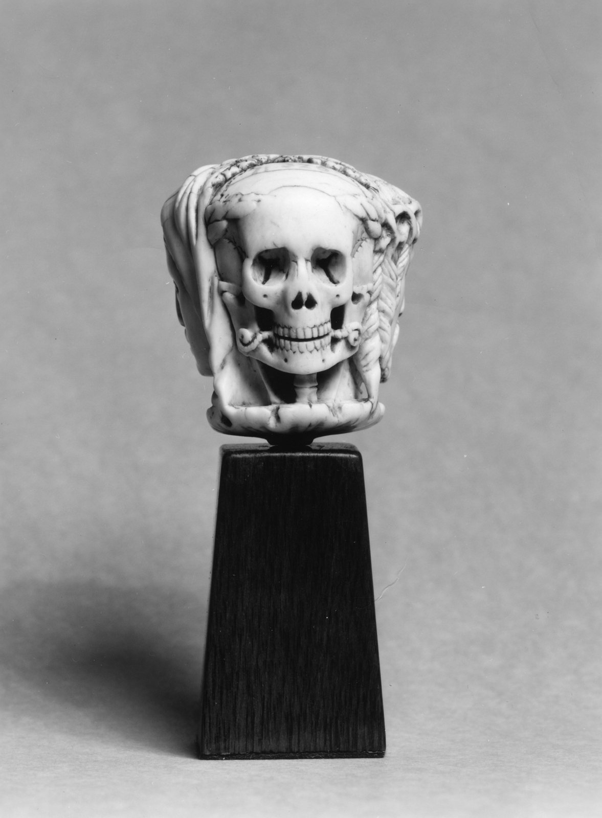 A small carved pendant on a black stand: a skull with a toothy grin sits at the front, with faces of Christ and the Virgin carved behind it