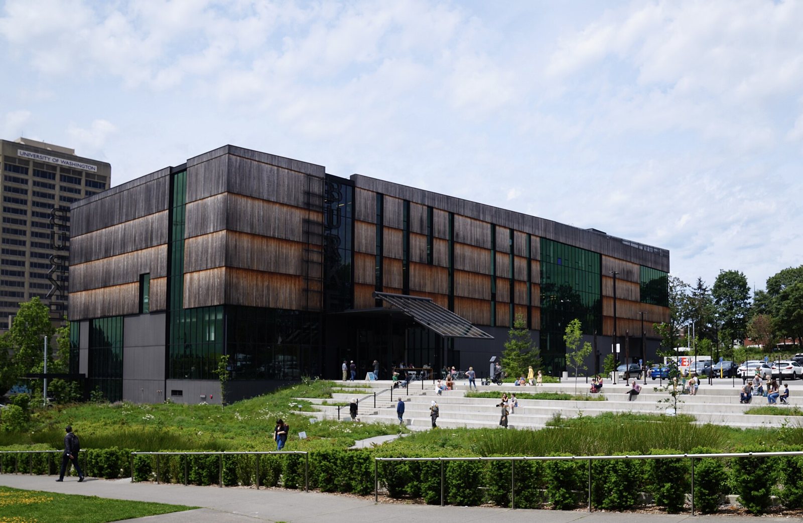 The Burke Museum in Seattle, a modern building clad in dark and amber wooden slats with large glass windows, with a stepped plaza and visitors in the foreground