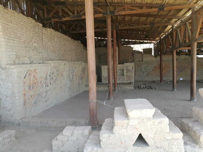 Bajo un techo de madera en la Huaca Cao Viejo, un largo muro de adobe pintado con franjas desvaídas en rojo, blanco y negro se alza sobre plataformas escalonadas bajas