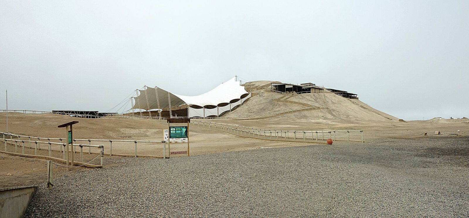 Vista amplia de la Huaca Cao Viejo en El Brujo, Perú: un gran techo blanco tensado protege el templo excavado sobre una colina arenosa, sobre un sendero de grava para visitantes