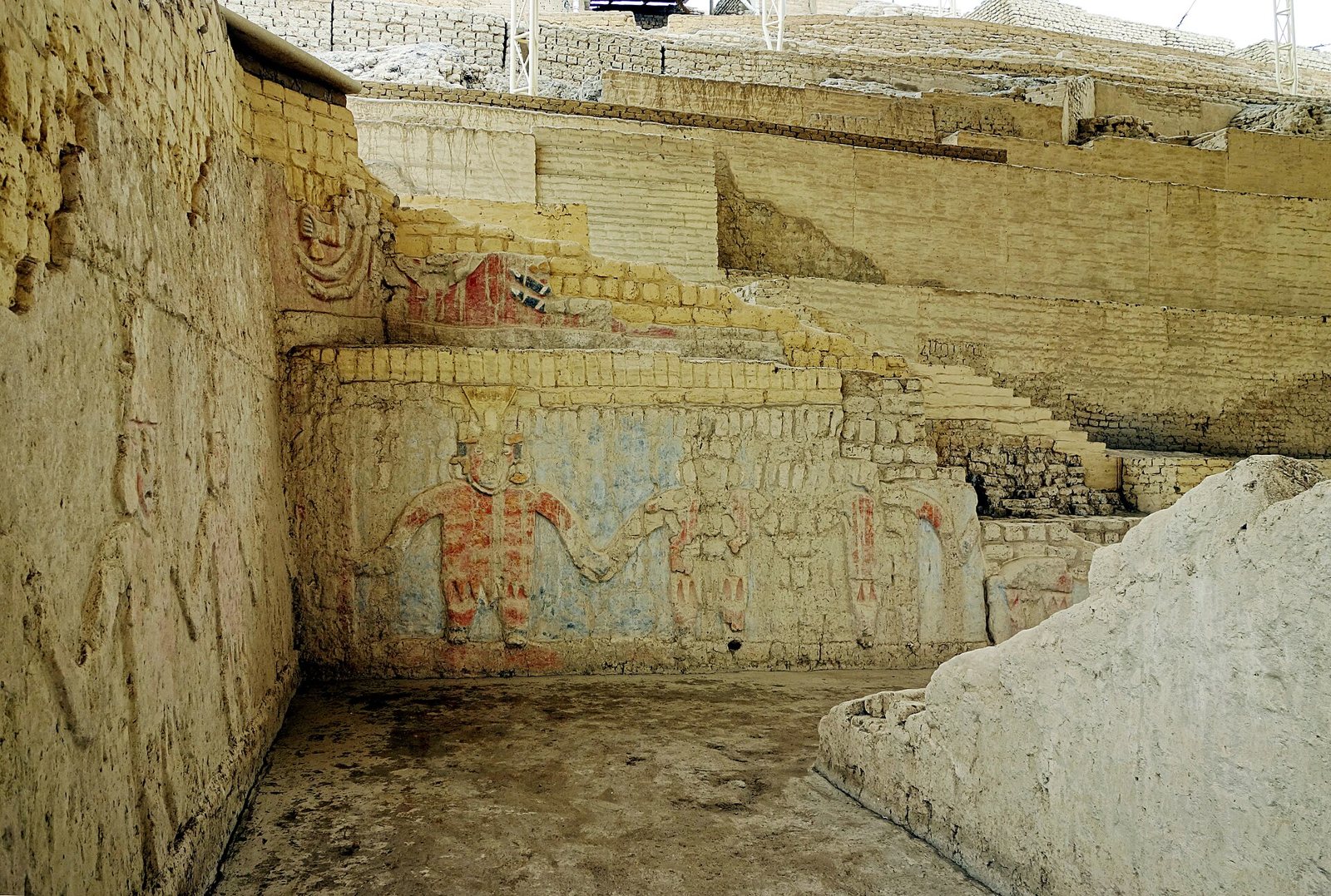 Un mural en relieve pintado sobre un muro de adobe escalonado en la Huaca Cao Viejo, con una fila de figuras rojas y ocres que se dan la mano