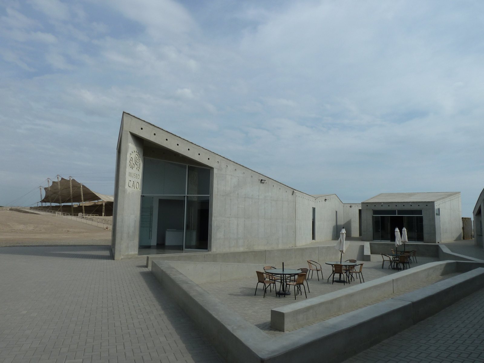 El Museo Cao en El Brujo: un edificio moderno de concreto con un frente de vidrio en ángulo y una terraza pavimentada con mesas, y al fondo la Huaca Cao Viejo con su techo de carpa