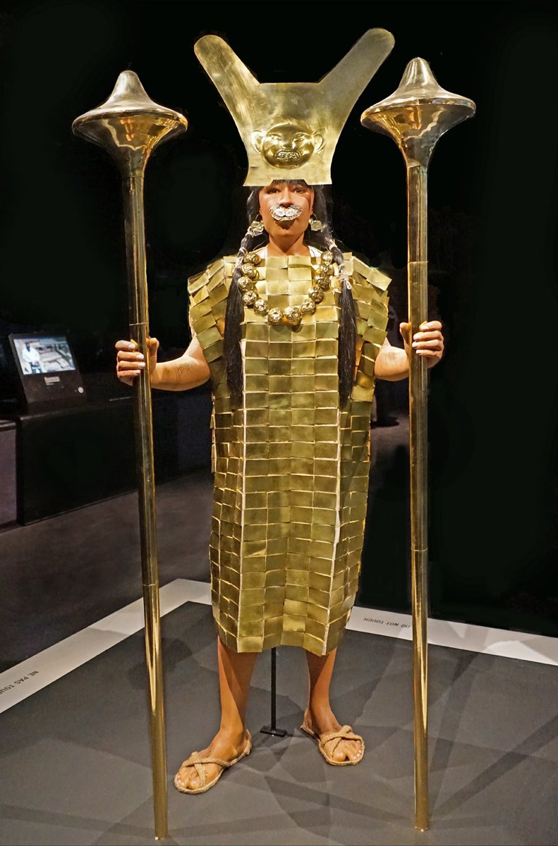 Una figura de museo de una mujer moche sobre un fondo oscuro, con una corona dorada, una túnica dorada y adornos, que sostiene una larga porra dorada en cada mano