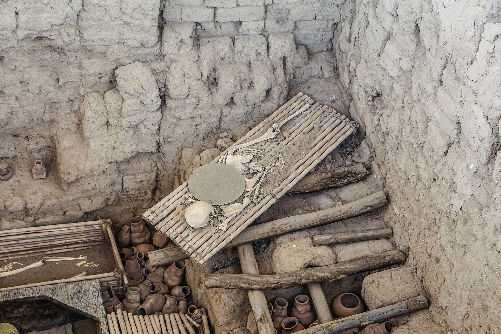 Dentro de una cámara de adobe claro, una plataforma de listones de madera se inclina sobre vasijas de cerámica, con huesos visibles sobre la plataforma y en un hueco abajo a la izquierda