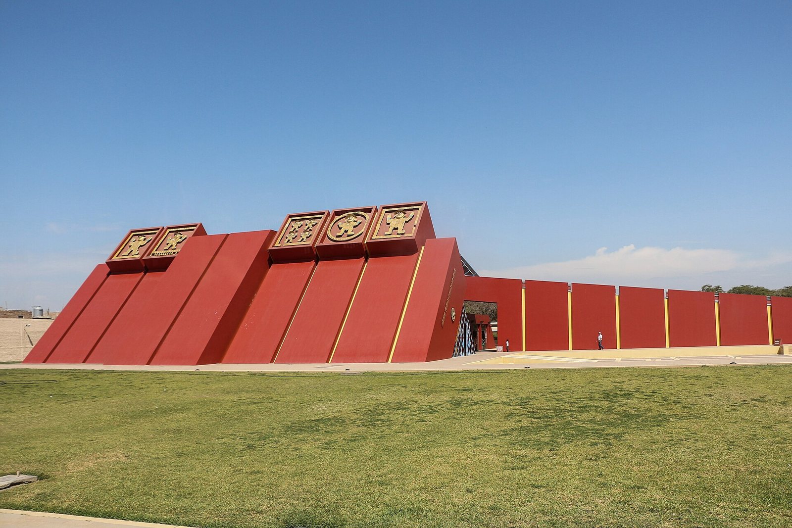 El Museo Tumbas Reales de Sipán, un edificio largo y rojo de muros inclinados en forma de pirámide, con placas doradas en el borde del techo, sobre un césped verde