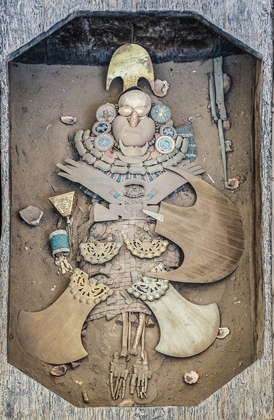 Vista desde arriba, una reconstrucción del entierro del Señor de Sipán: una figura con un tocado de media luna dorado yace en un marco de madera entre adornos de oro, turquesa y concha, con grandes medias lunas doradas a los lados