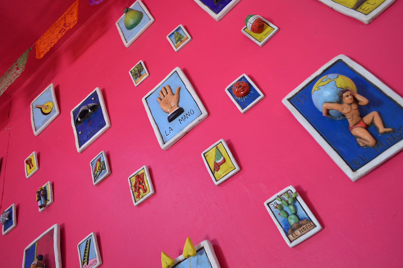 Handmade clay lotería tiles mounted at angles on a bright pink wall, among them La Mano, a cactus and a man holding up a globe, each in a white frame