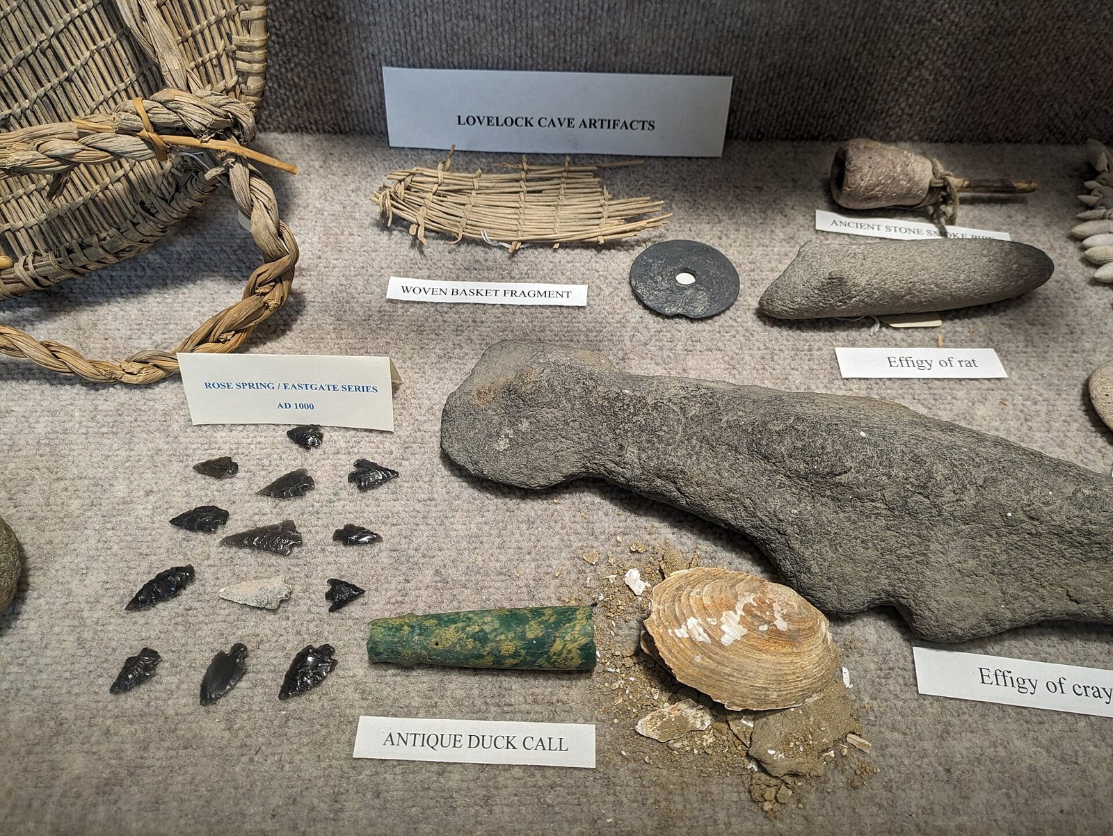 A museum case of Lovelock Cave objects with paper labels: a coiled basket at the top left, a basket fragment, dark stone arrow points, a green bead, a shell and a carved stone effigy