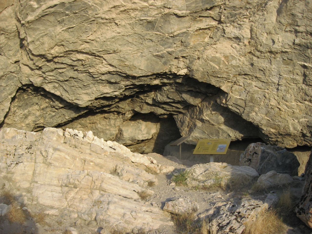 The mouth of Lovelock Cave: a low, dark opening under a large overhang of yellow-brown rock, with a small yellow information sign among the rocks at the lower right