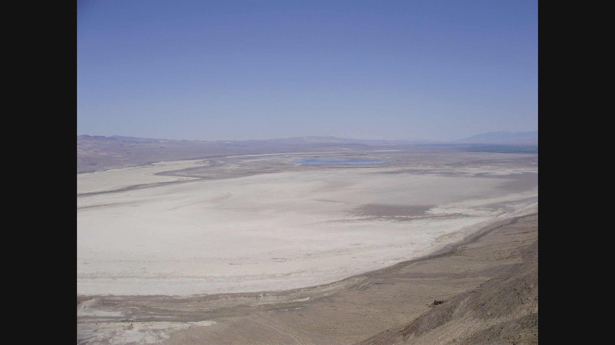 A wide view of the Humboldt Sink in Nevada from a hilltop: a huge pale, flat dry lake bed with a small patch of water, brown hills along the edge and a hazy sky
