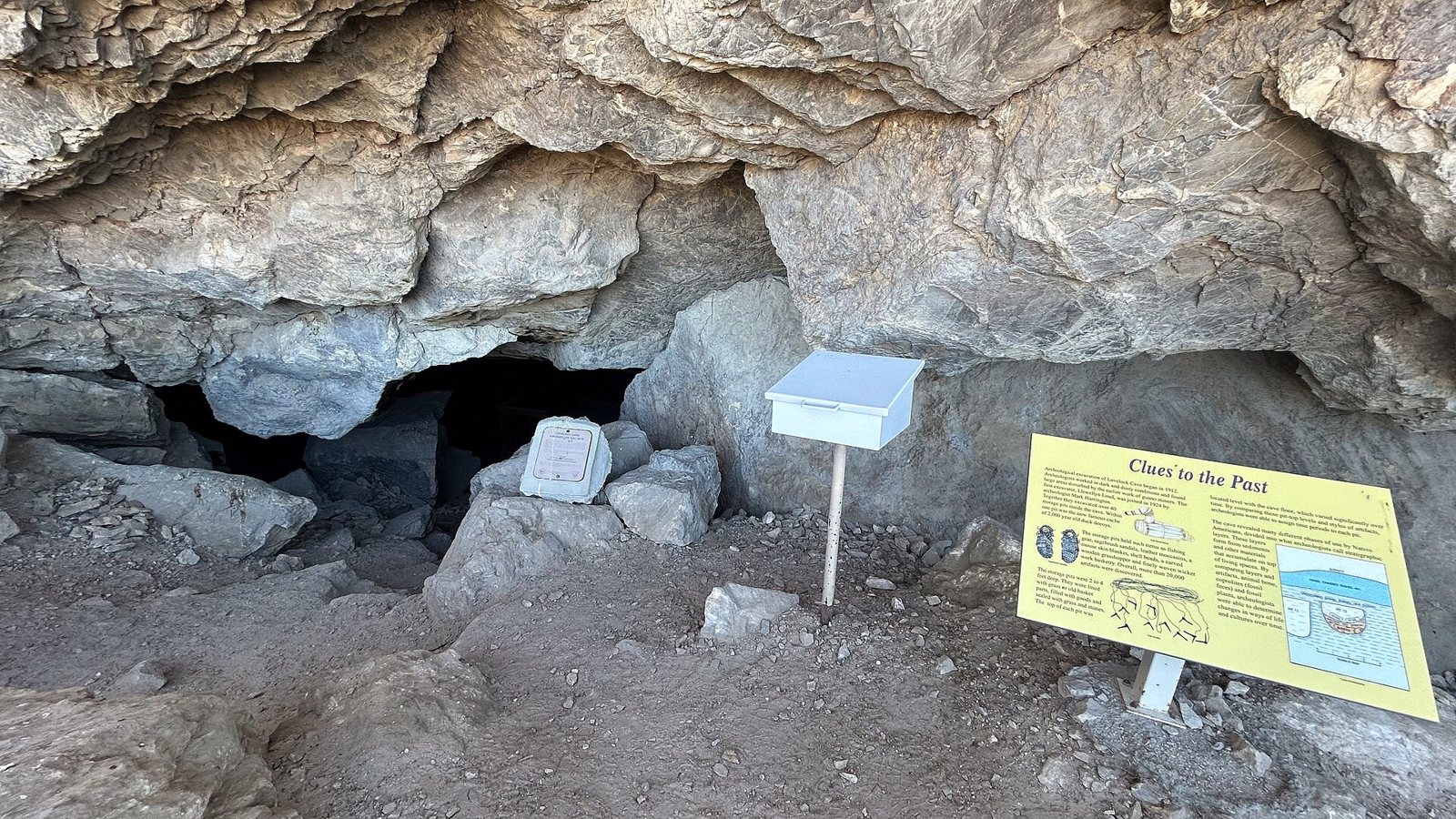 The entrance of Lovelock Cave beneath a rock overhang, with a white registry box on a post and a yellow information board in front of the dark opening
