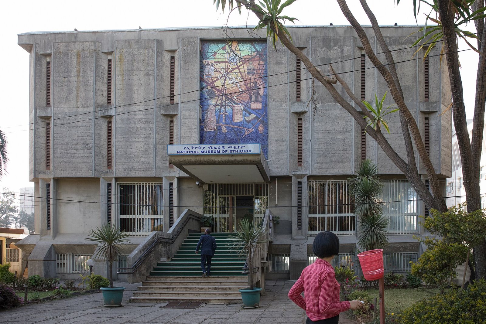 Concrete front of the National Museum of Ethiopia in Addis Ababa, with a large mosaic panel above the entrance, a flight of steps, potted palms and two visitors outside