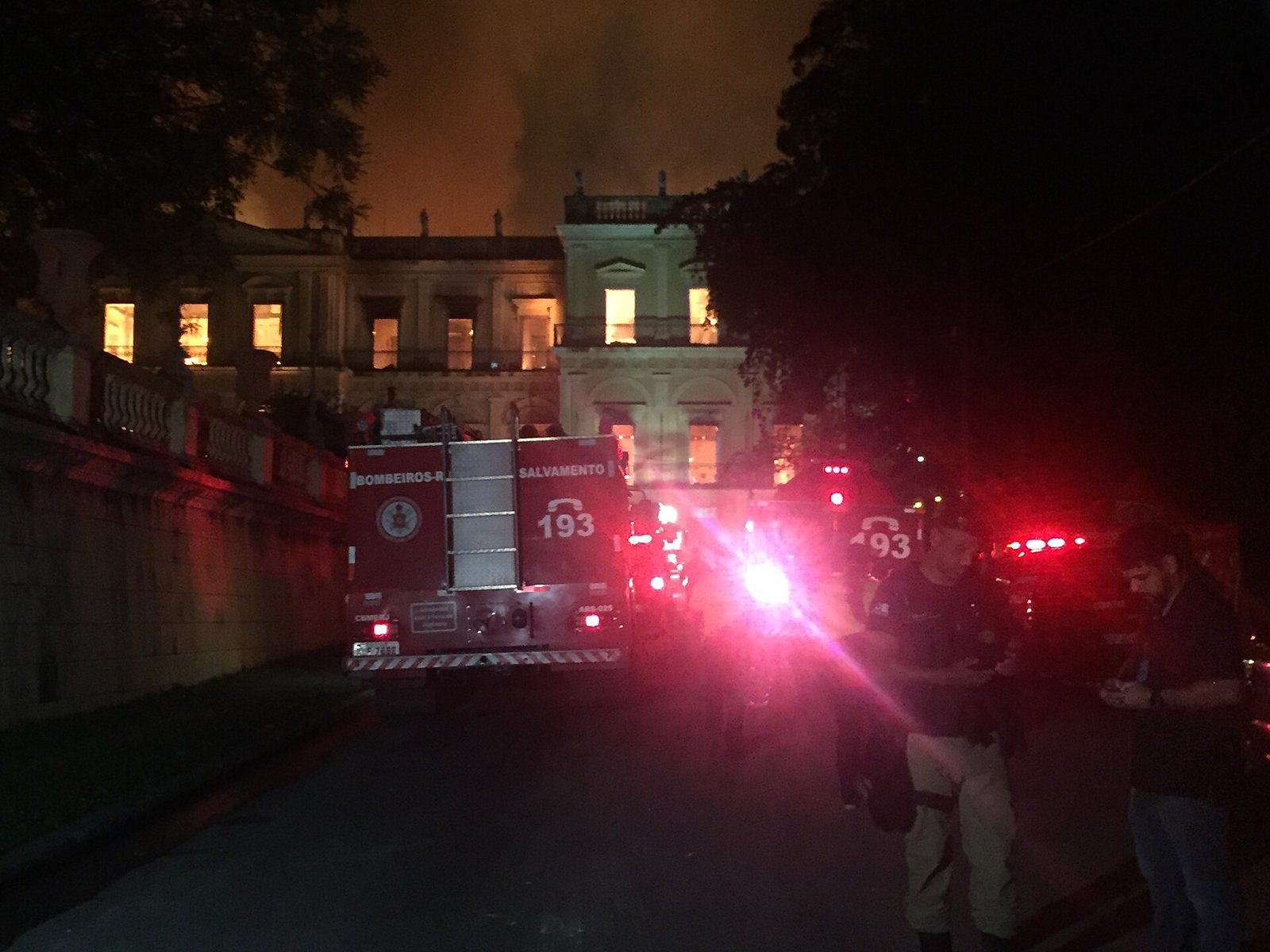 The Museu Nacional building at night with fire glowing behind its windows, a fire engine and firefighters in the foreground and red emergency lights