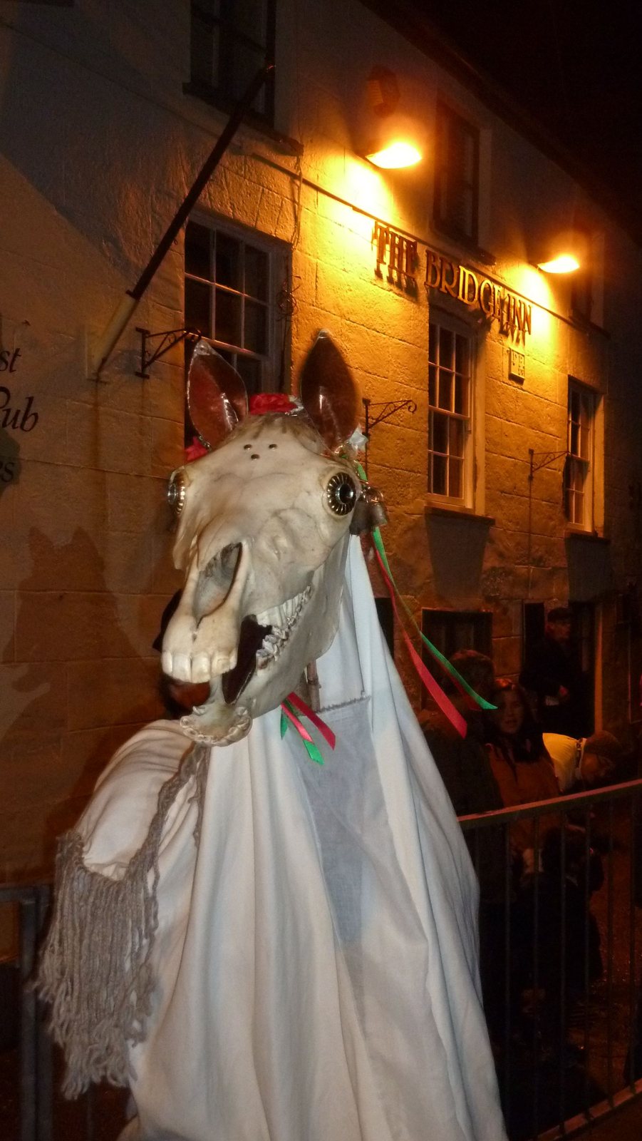 A Mari Lwyd at night outside the Bridge Inn in Chepstow: a horse skull draped in a white sheet under the glow of the pub's lamps and sign