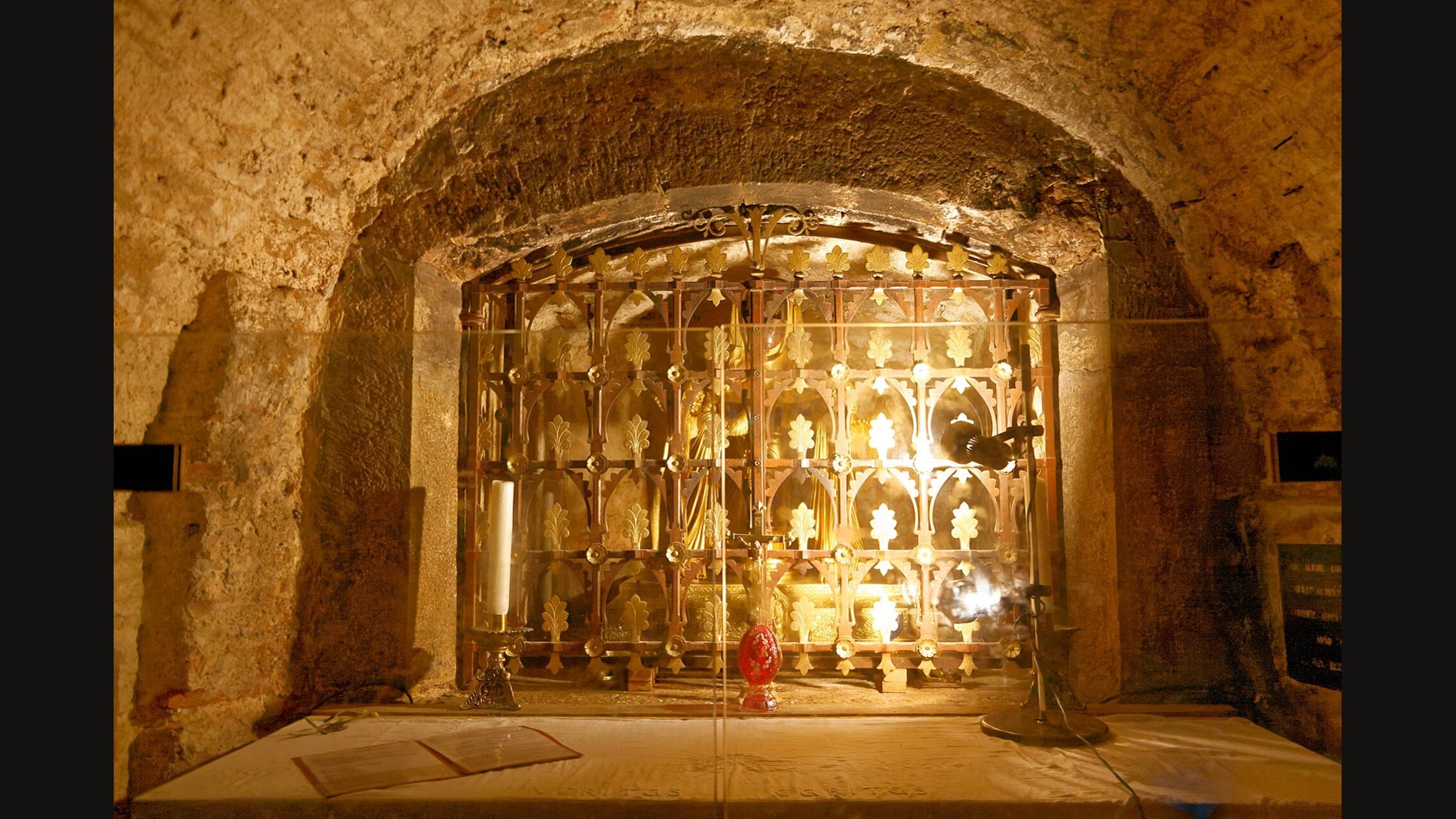 The reliquary shrine in the crypt of the Saint-Maximin basilica: an ornate gilded grille set in an arched niche of rough stone, glowing in the lamplight