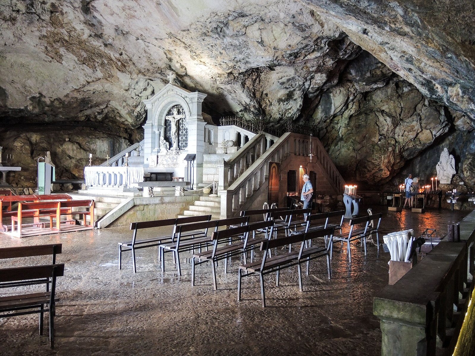 Inside the Sainte-Baume cave sanctuary: a white altar with a crucifix under the rock ceiling, with rows of wooden benches and a few visitors