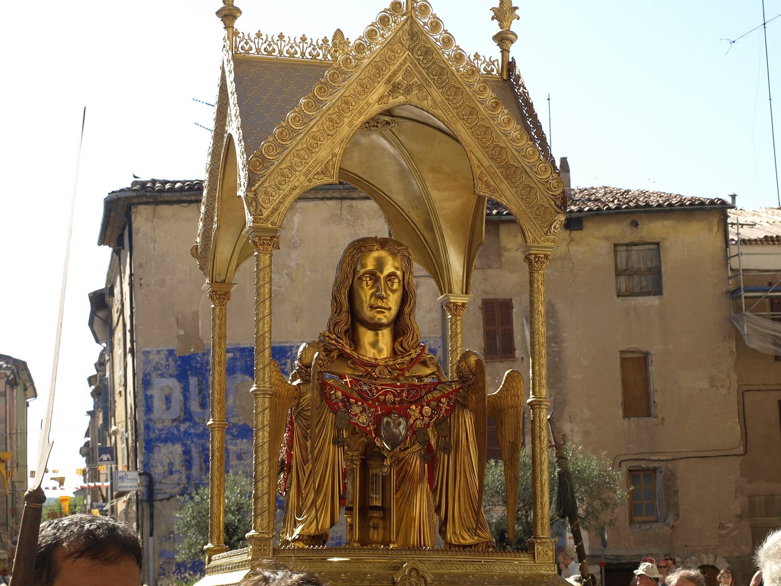 A gilded bust reliquary under a small golden canopy, carried through a street in front of old stone houses