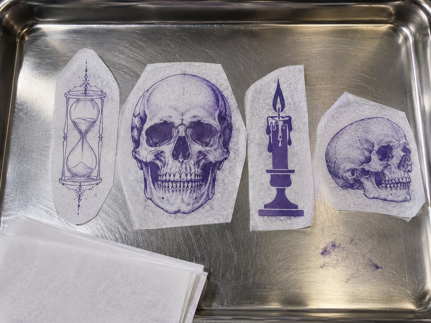 Illustration: Row of purple tattoo transfer stencils on a steel tray: a fine-line hourglass, a bold traditional skull, a blackwork candle and a small engraving-style skull, beside blank stencil paper