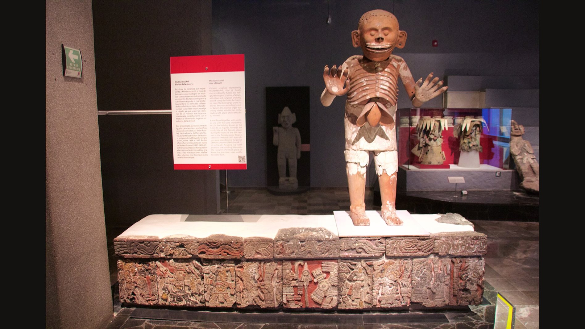 Life-size clay statue of Mictlantecuhtli in a museum gallery: a skeletal figure with a grinning skull face, rows of exposed ribs, a hanging organ below the ribs and both clawed hands raised, standing on a carved stone base beside a label panel