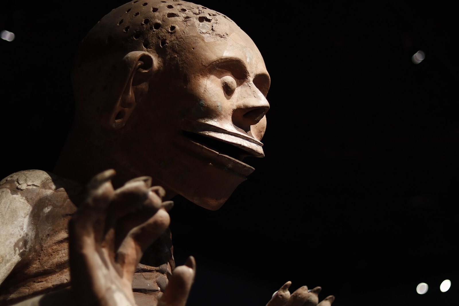 Close-up of the clay Mictlantecuhtli statue against a dark background: a skull-like head pierced with small holes, an open lipless mouth and a clawed hand raised in front
