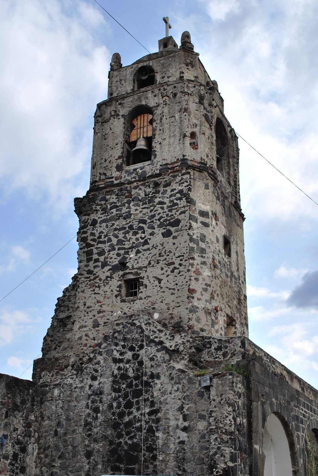 Una torre de campanas de piedra oscura de la iglesia original de Mixquic, contra un cielo azul con nubes, con una campana visible en la abertura superior