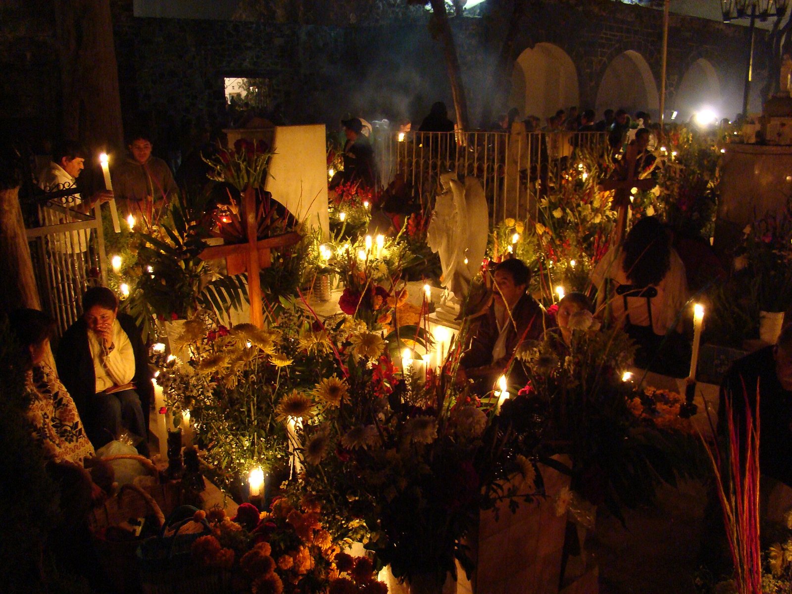 Familias sentadas entre tumbas cubiertas de flores en el panteón de Mixquic de noche, iluminadas por muchas velas y con una cruz de madera al centro
