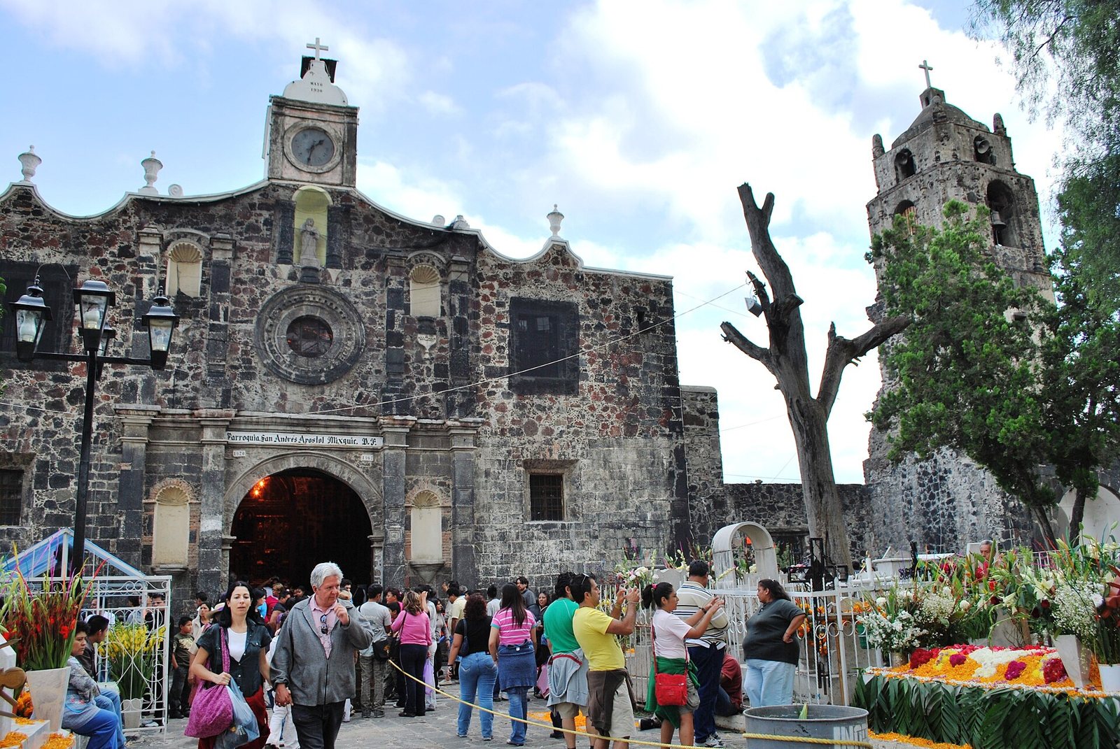 La iglesia de piedra de San Andrés Apóstol en Mixquic, con un reloj sobre la puerta de arco, una torre de campanas a la derecha y visitantes reunidos en el atrio