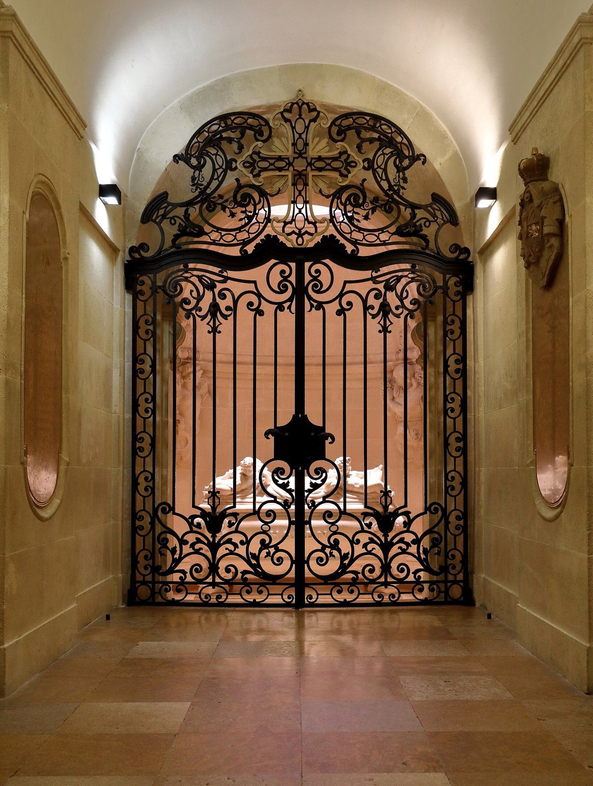 A wrought-iron gate with a cross at its crest, closing a lit stone chamber where a white marble tomb with carved figures stands