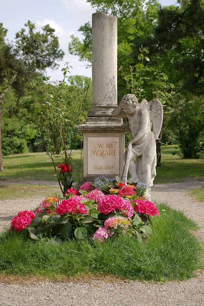 A stone memorial in a park: a broken column on a pedestal inscribed W. A. Mozart 1756 to 1791, with a mourning angel statue leaning beside it and pink hydrangeas at its base