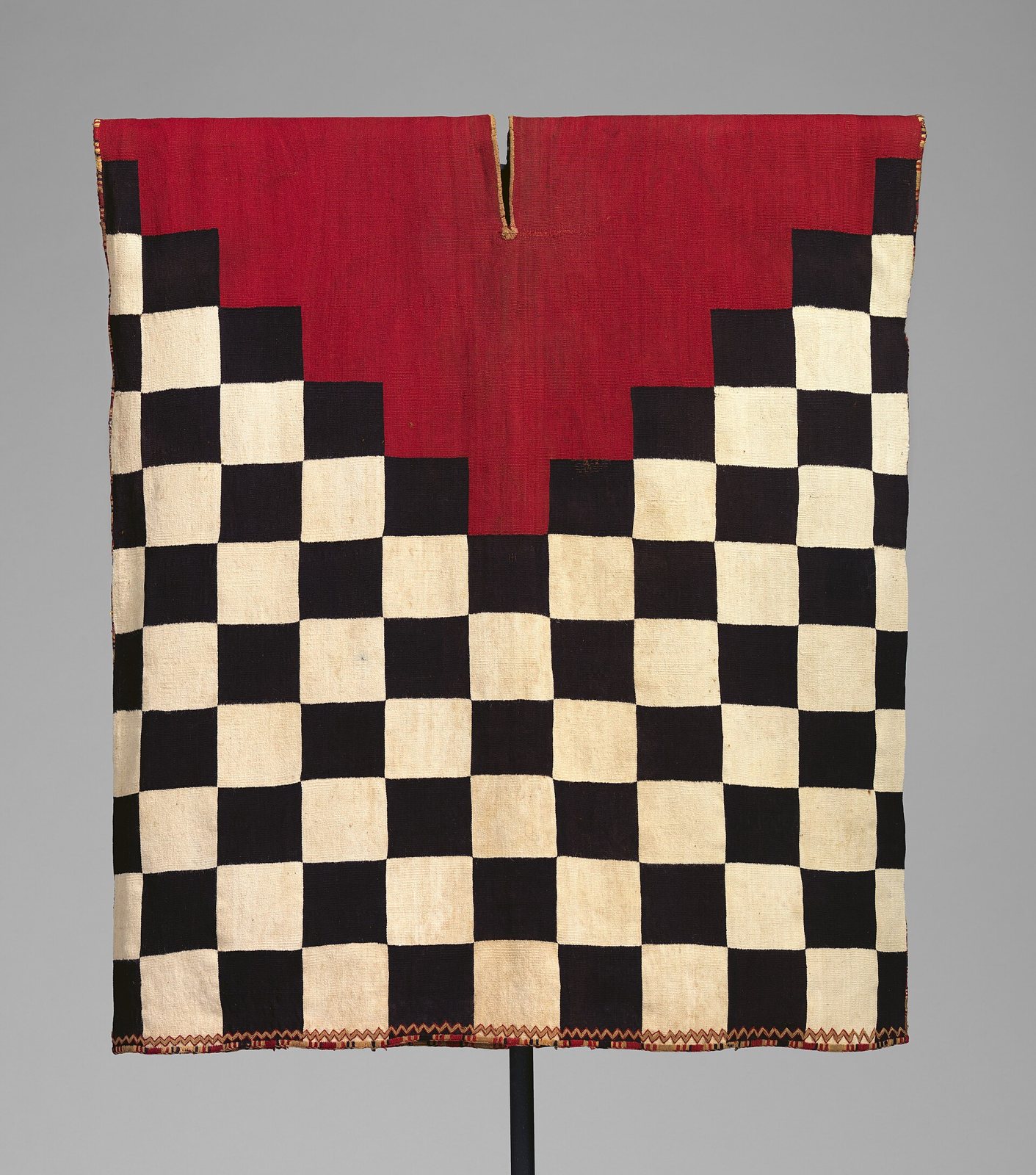An Inca tapestry-woven tunic with a red V-shaped yoke above a black-and-white checkerboard body, hung on a stand against a grey background
