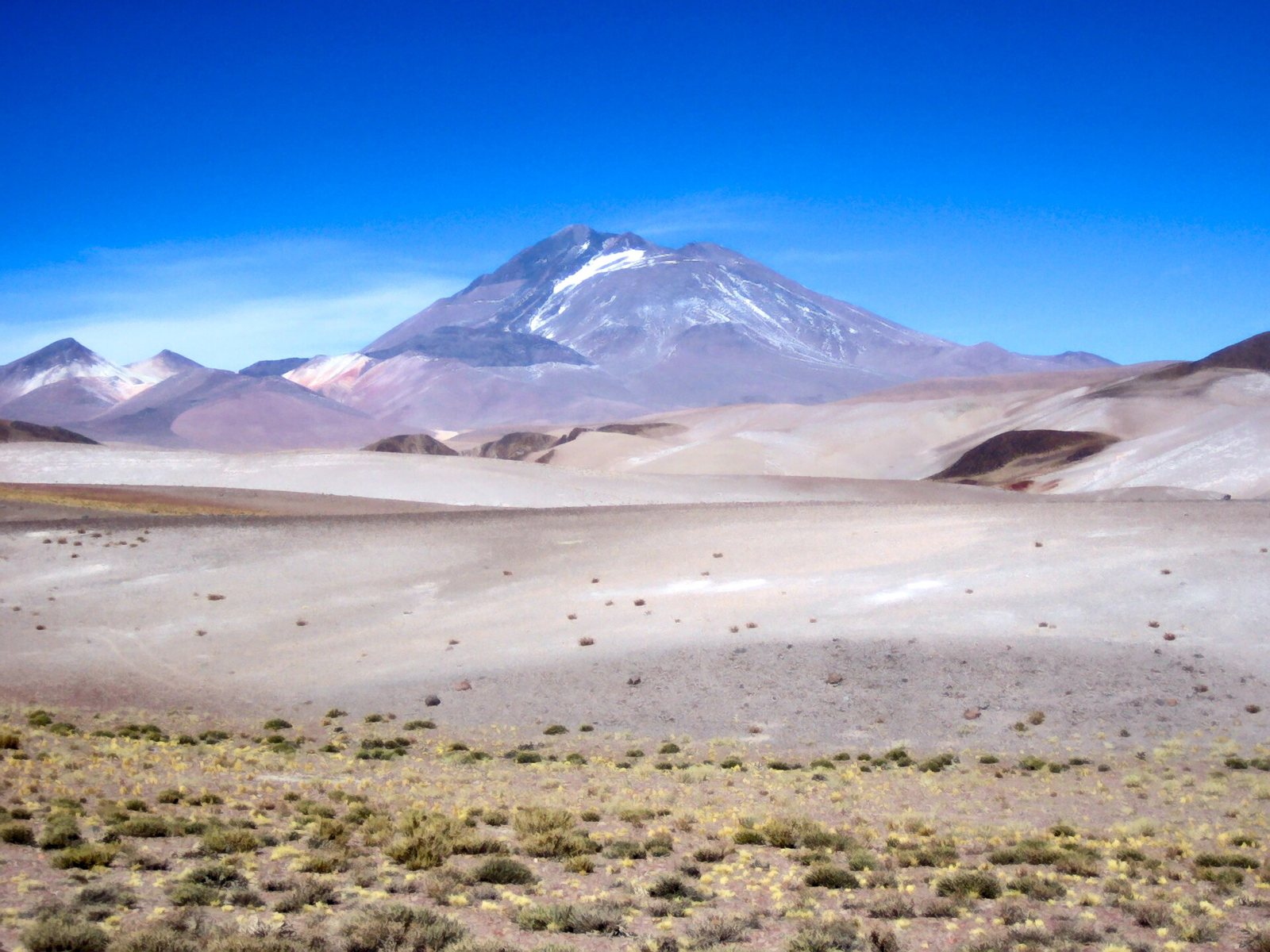 Llullaillaco volcano in Salta Province, Argentina: a dry, snow-streaked mountain beyond a pale desert plain dotted with low shrubs, under a deep blue sky