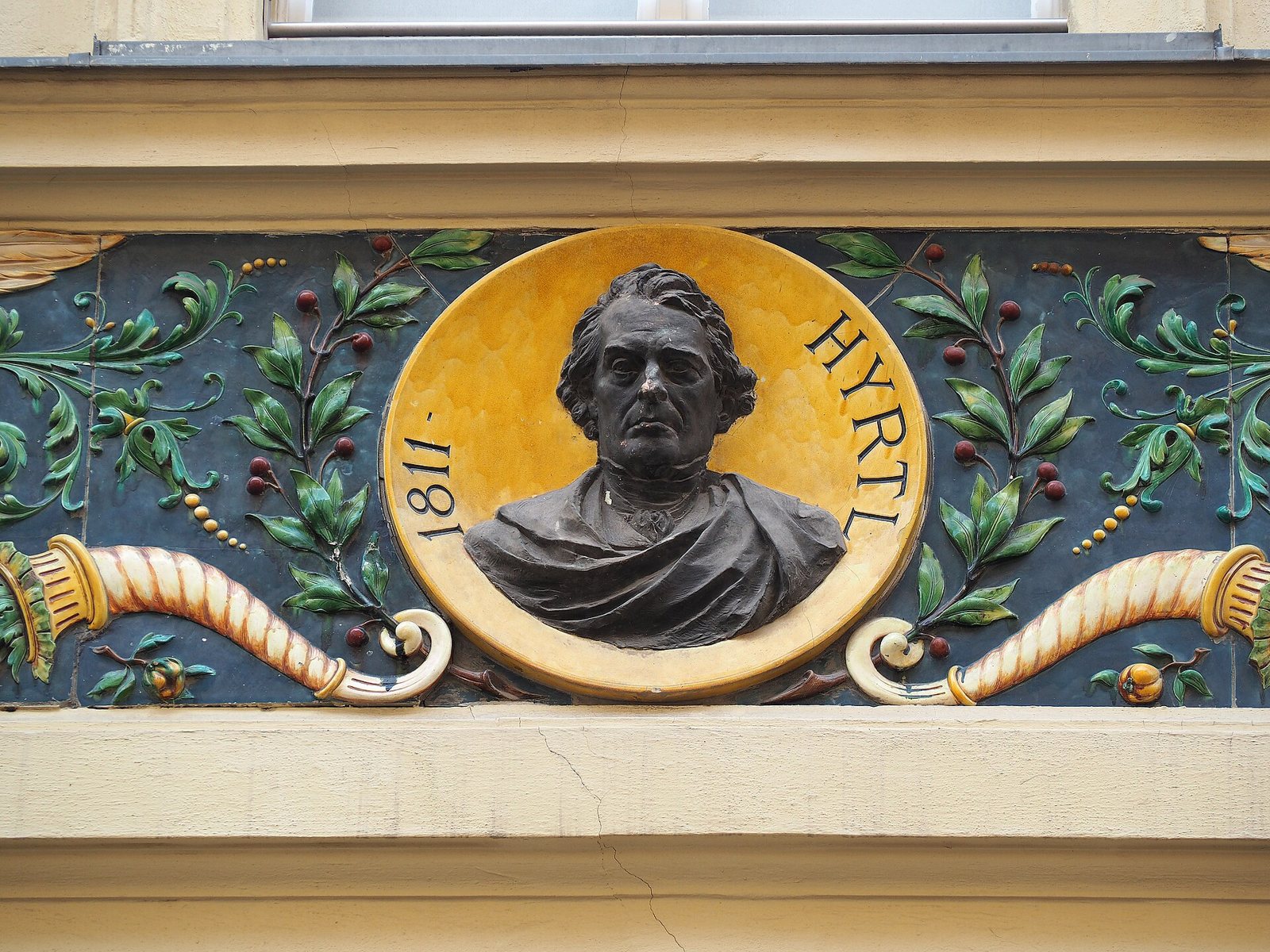 A dark bronze bust of Joseph Hyrtl in a gold circle lettered Hyrtl and 1811, set in a colored tile relief with green leaves, red berries and cornucopias on a blue ground