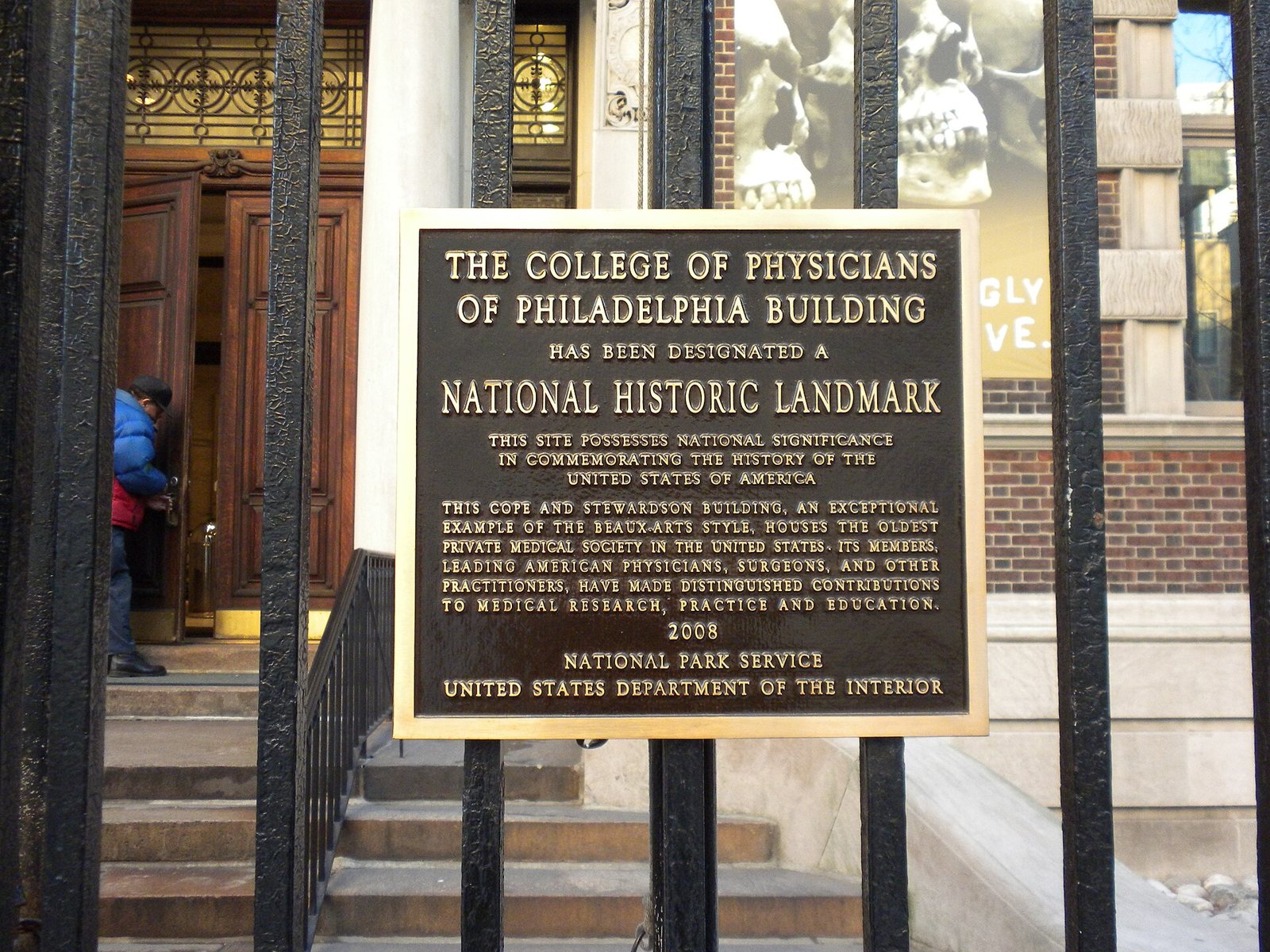 A bronze plaque on an iron fence reading The College of Physicians of Philadelphia Building has been designated a National Historic Landmark, with wooden doors and steps behind it