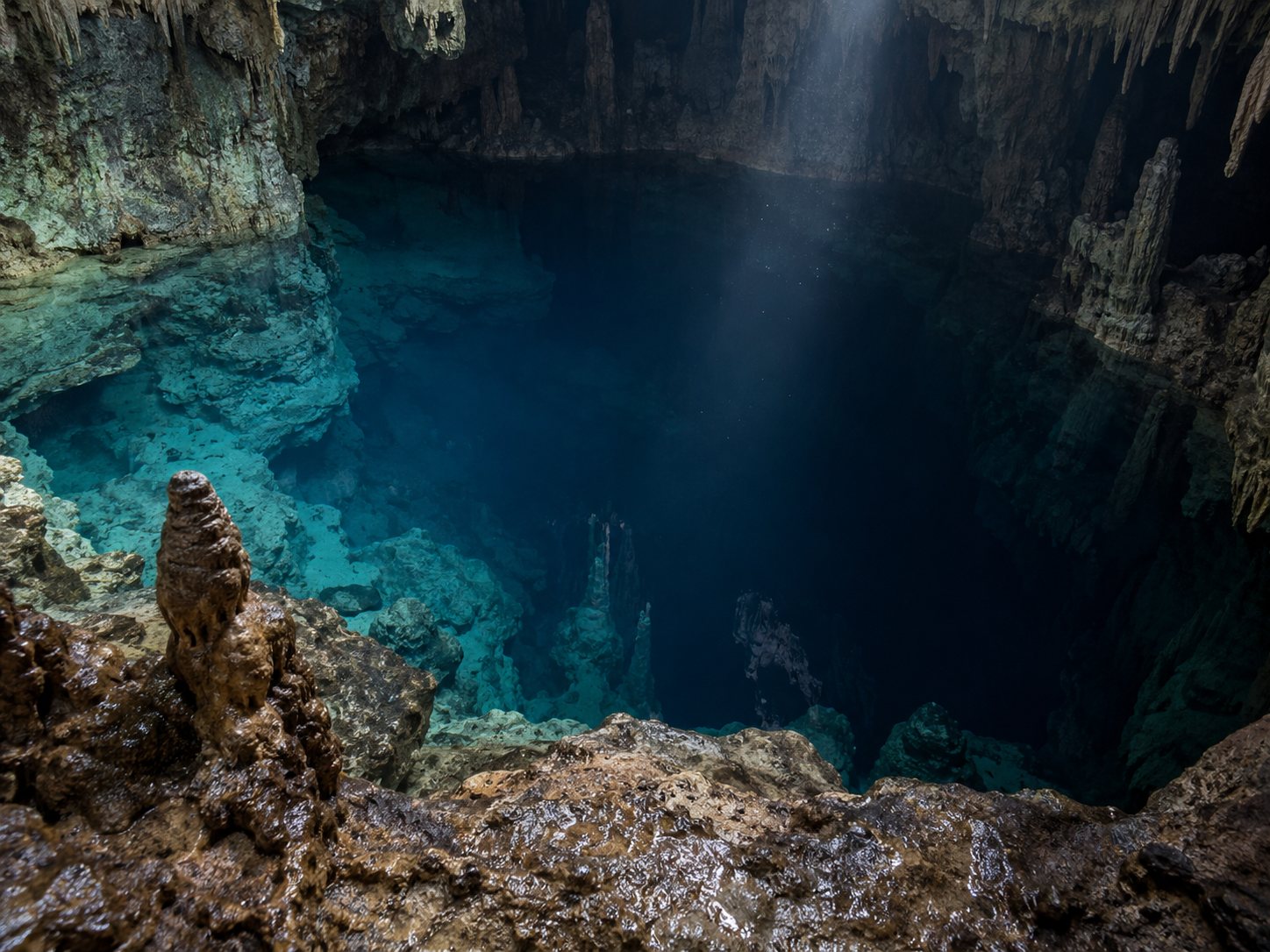 Illustration: View down into a deep flooded cave pit with clear water fading from pale blue to near black and limestone walls with stalactites and stalagmites in a distant light beam