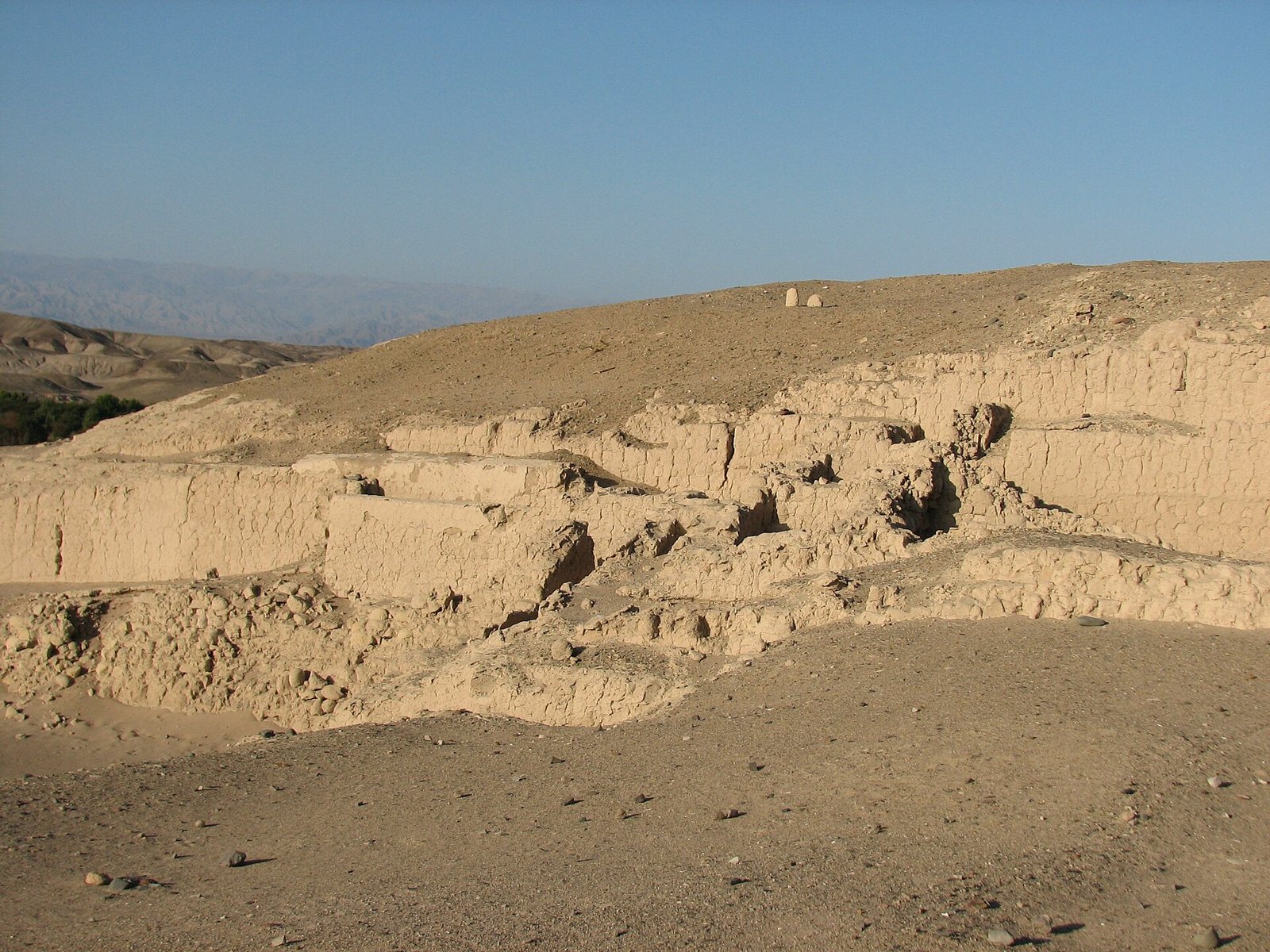 Eroded adobe walls and stepped terraces of the Cahuachi ruins on a sandy hillside under a clear blue sky, with dry mountains far behind
