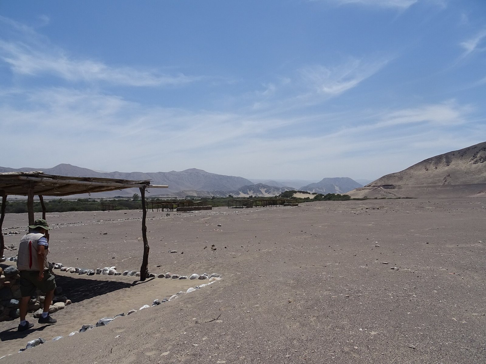 The Chauchilla cemetery site in the Peruvian desert: a wide gravel plain with a rope-and-stone path, a wooden shelter on the left, a visitor standing beneath it and dry mountains behind