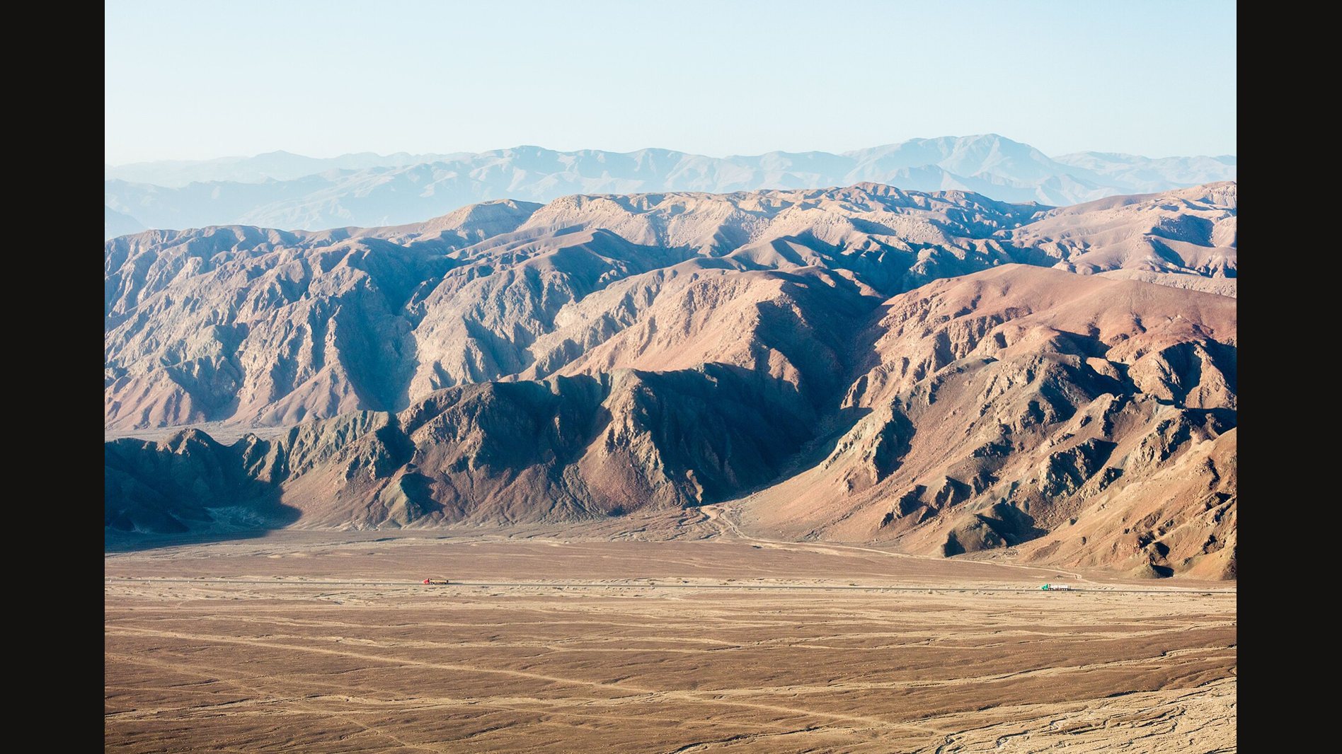 The dry Nazca plain in southern Peru seen from a high viewpoint: a flat tan desert floor with faint tracks across it, below rust-brown ridges and a hazy line of mountains
