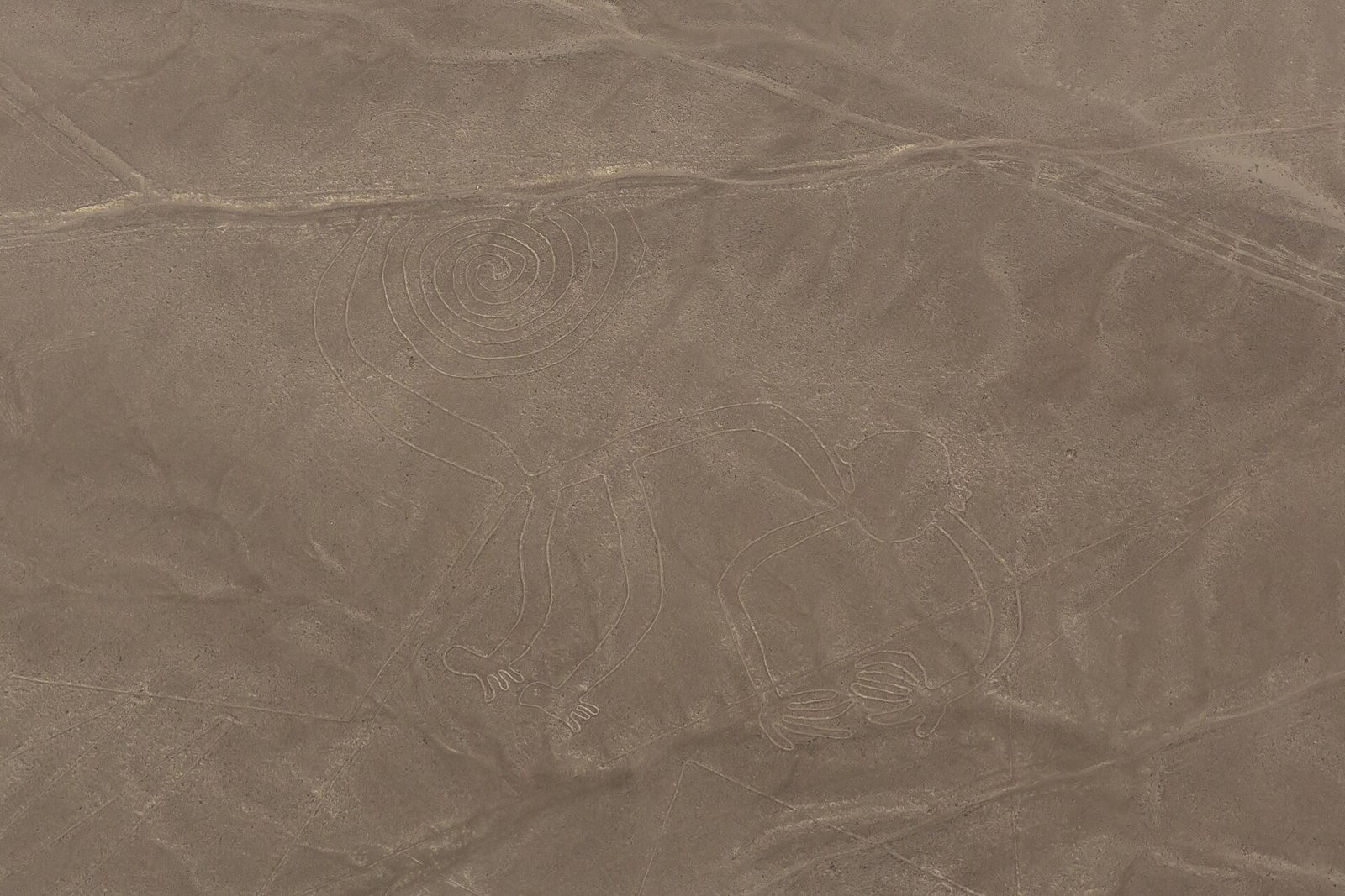 A flat, pale brown stretch of desert photographed from the air, with thin pale lines scratched into the ground and a spiral shape near the center