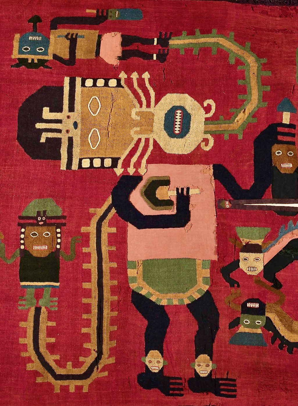 A detail of a red Nasca textile with dark outlined figures in colorful headdresses and masks, each joined by a looping serpent-like band to smaller figures
