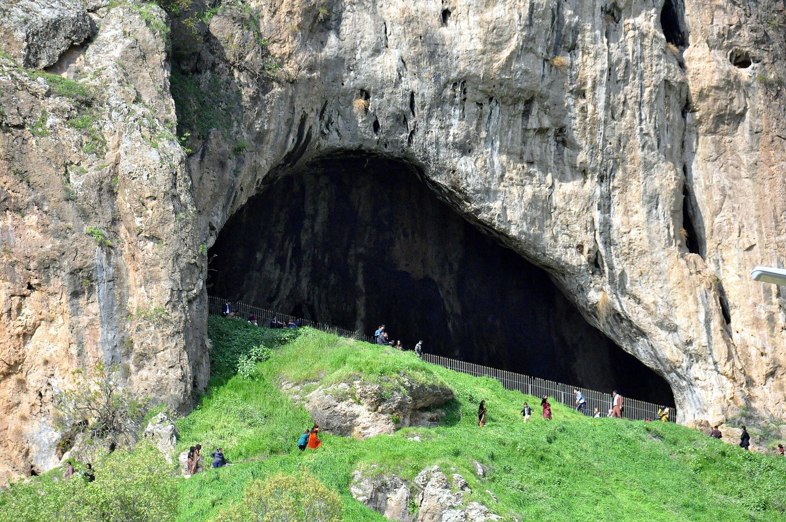 The wide arched mouth of Shanidar Cave, a dark cavern in a pale limestone cliff above a steep grassy slope, with small groups of visitors along a fence