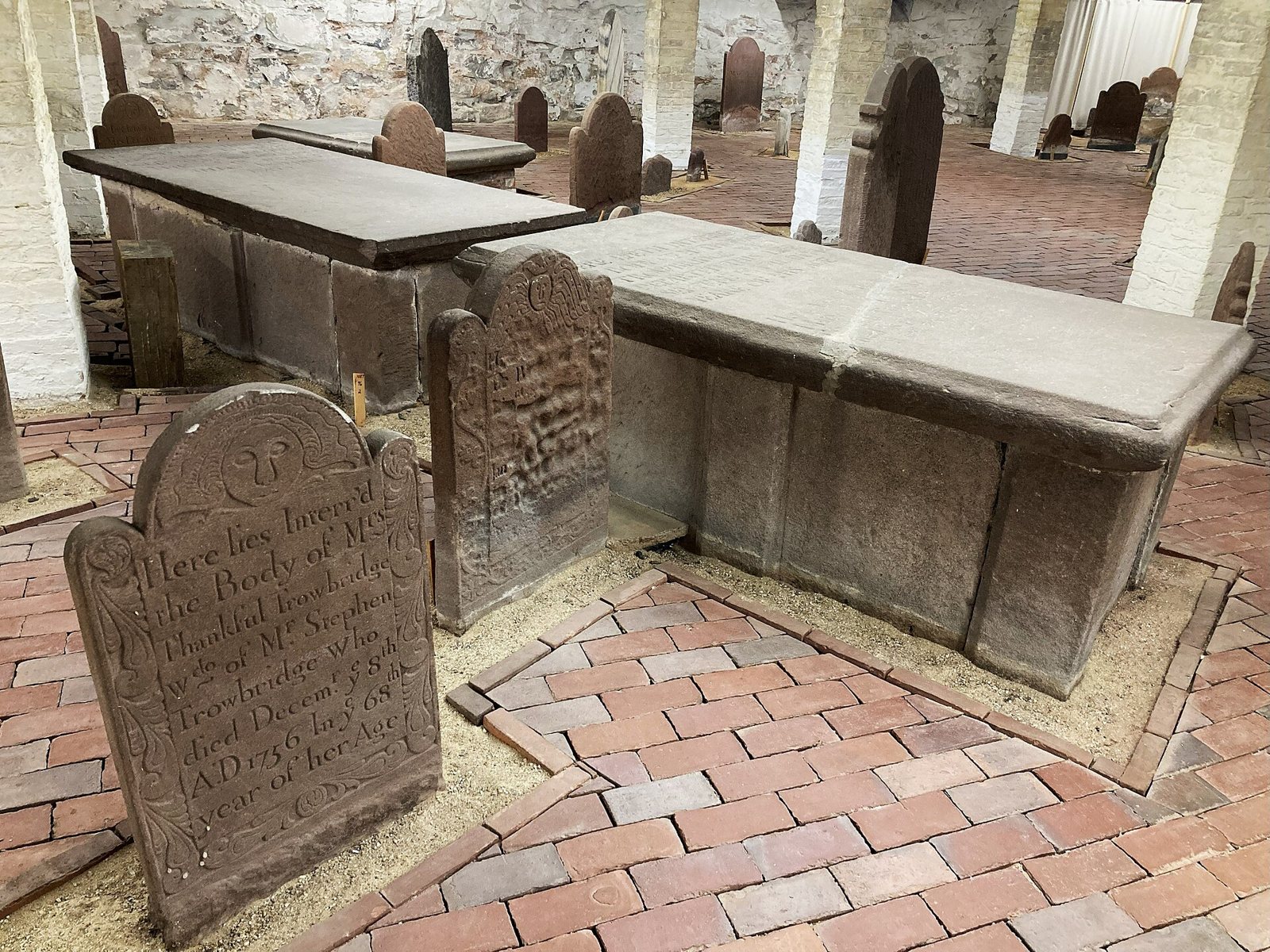 Old brownstone headstones and two large table tombs on a brick floor in the crypt beneath Center Church