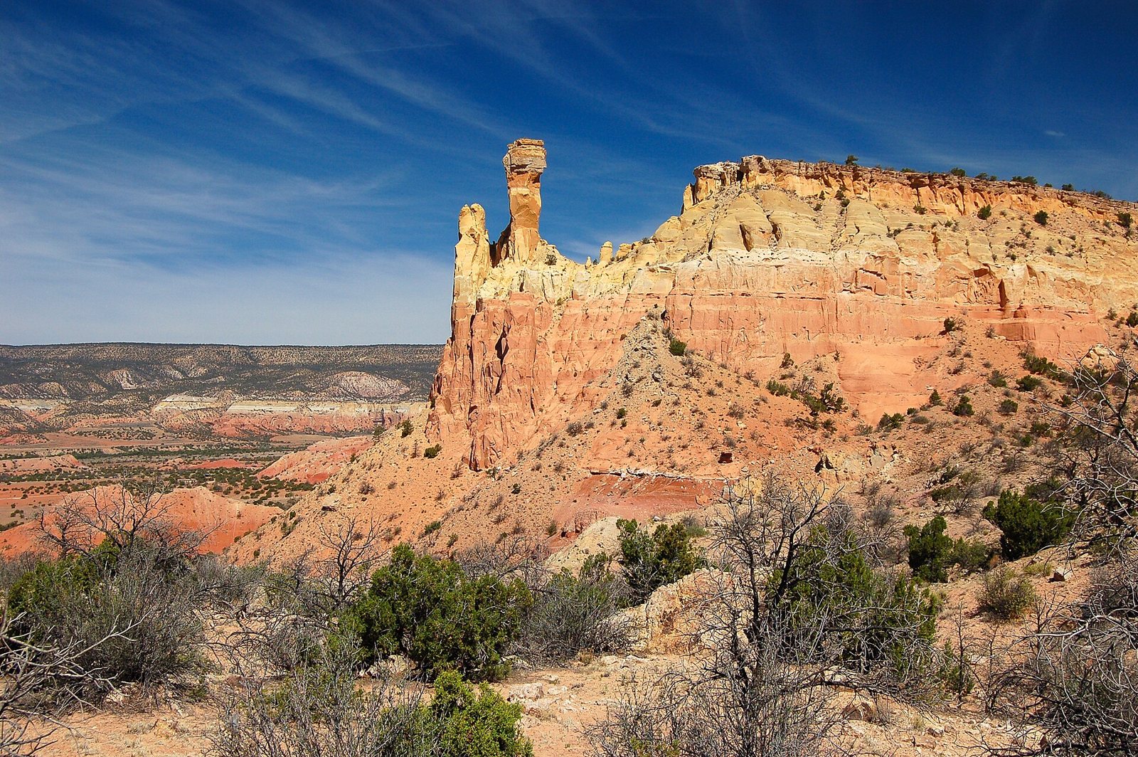 A tall red sandstone cliff at Ghost Ranch with a narrow chimney-like pinnacle on its edge, desert scrub in the foreground and blue sky above