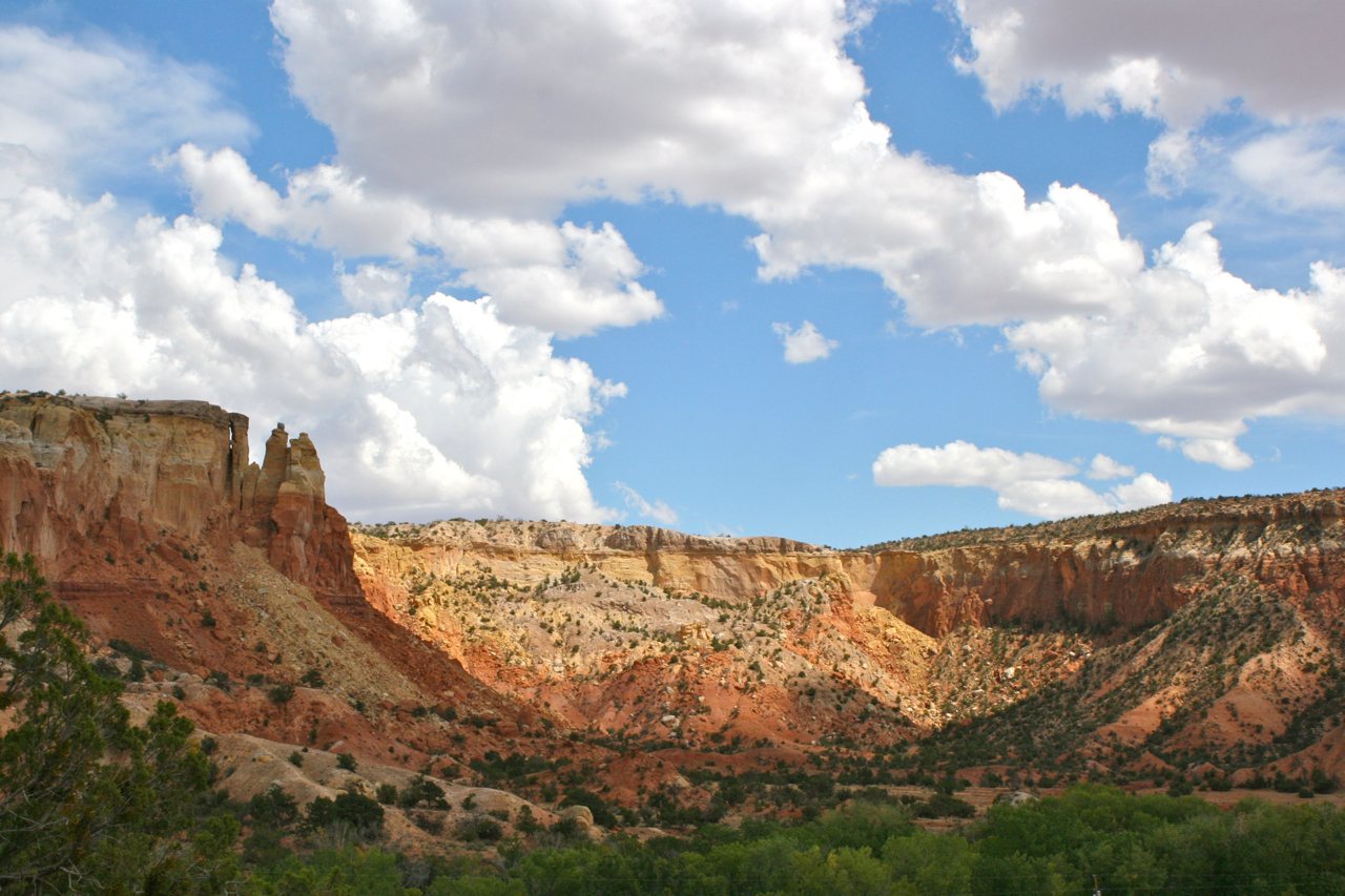 Red and orange sandstone cliffs and flat-topped mesas at Ghost Ranch, New Mexico, with green trees below and white clouds in a blue sky