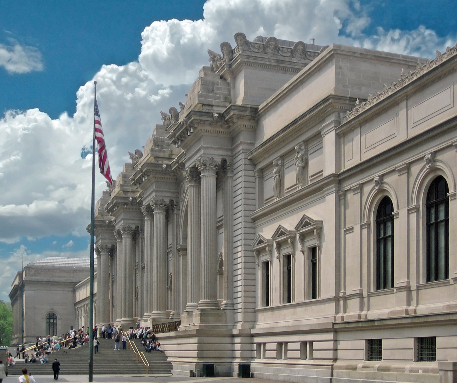 The pale stone Fifth Avenue facade of the Metropolitan Museum of Art, with paired columns, tall arched windows, a flag on a pole and visitors on the steps