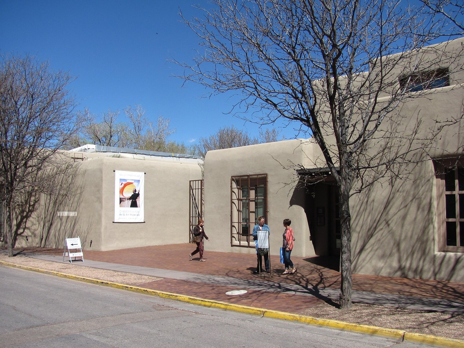 A low, flat-roofed adobe-style museum building in Santa Fe with a poster on its wall, bare trees and a few people on the sidewalk