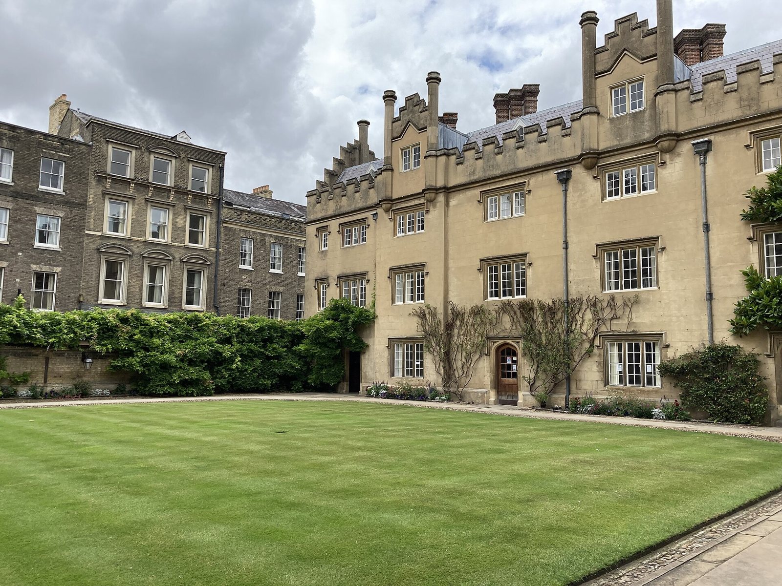 Hall Court at Sidney Sussex College, Cambridge: a mown lawn before a battlemented stone range with tall chimneys and climbing plants, and brick buildings to the left
