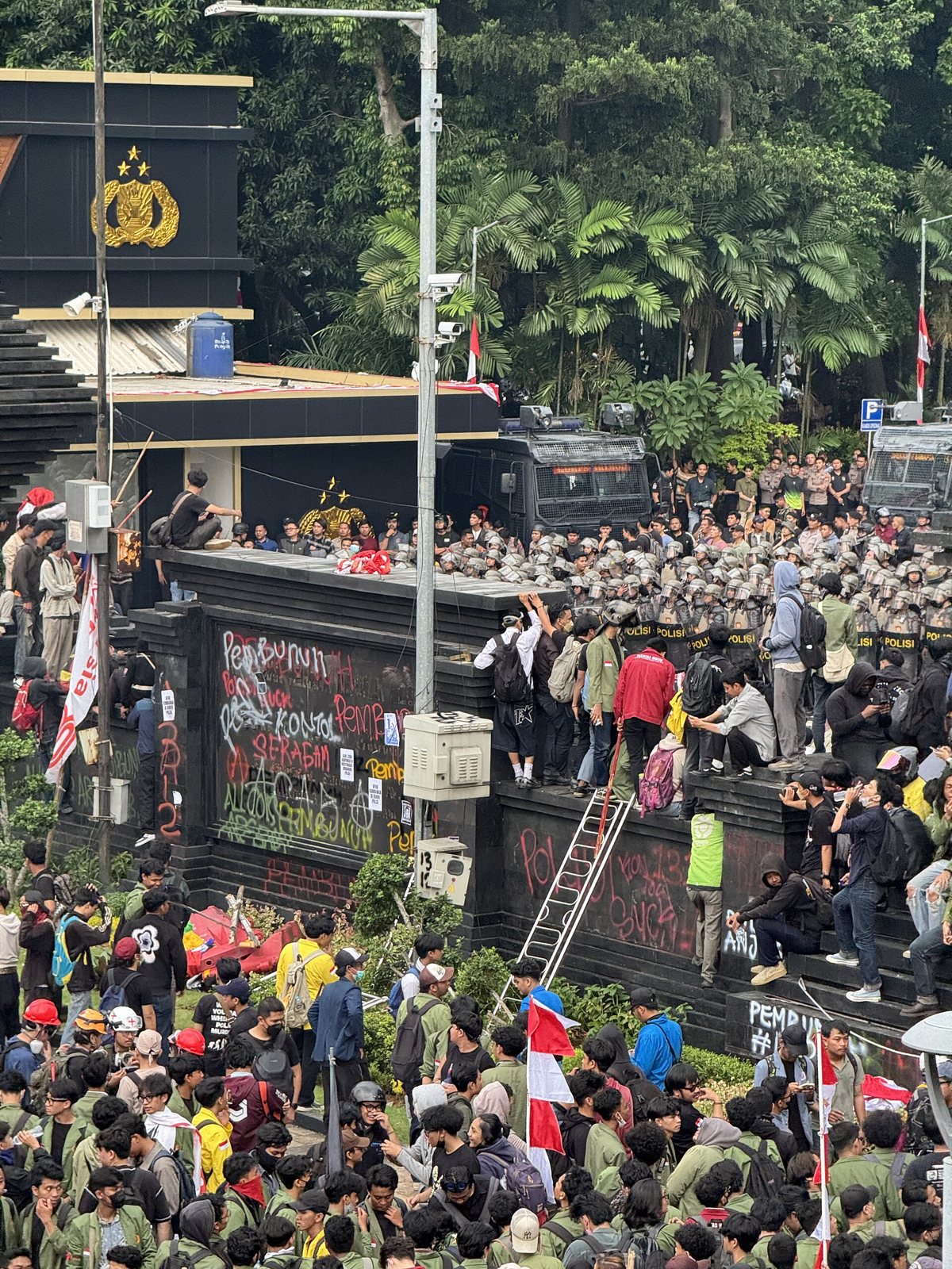 Estudiantes reunidos ante un portón negro coronado por un emblema policial en Yakarta, con filas de policías antidisturbios tras el muro y banderas rojas y blancas entre la multitud