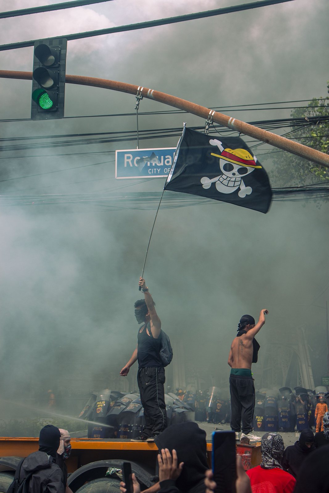 Un manifestante enmascarado, de pie sobre un vehículo, alza una bandera negra con una calavera con sombrero de paja sobre huesos cruzados, colgada de una farola entre mucho humo, con escudos antidisturbios abajo